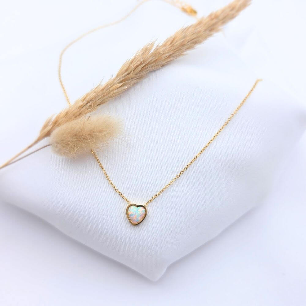 Collier chaîne dorée fine en acier inoxydable avec pendentif cœur en opale australienne blanche aux reflets irisés, présenté sur un tissu blanc avec un épi de blé séché.
