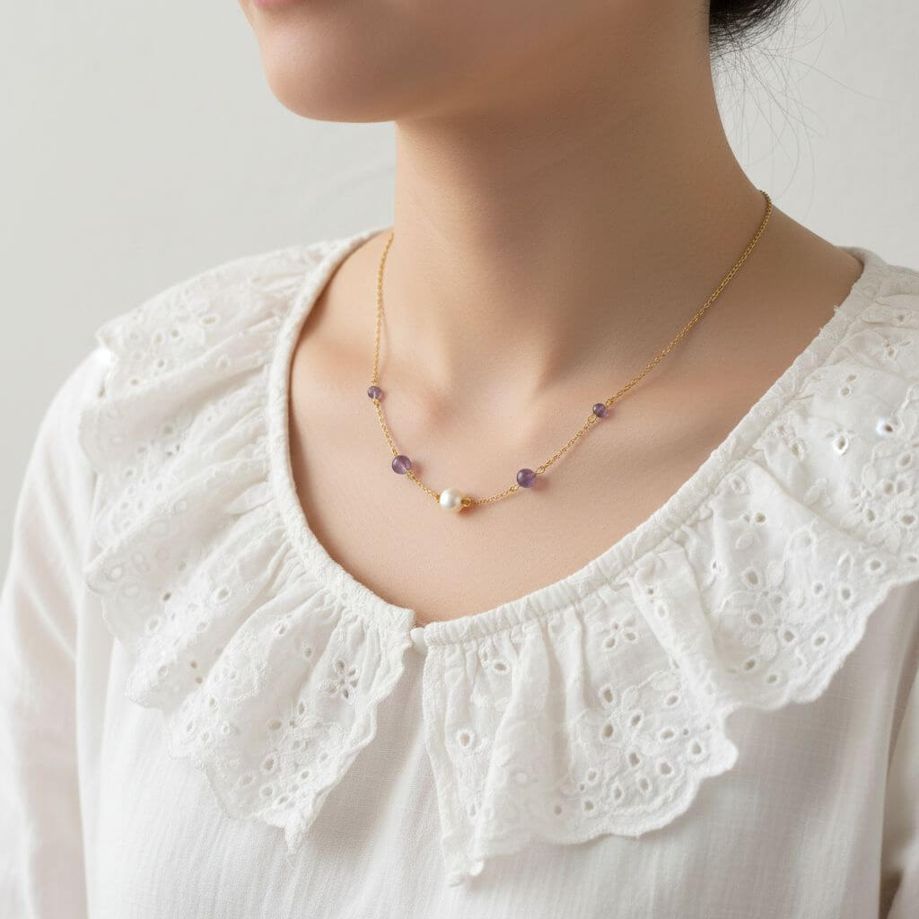 Gros plan sur une personne portant un collier fin en chaîne dorée. Le bijou comporte une perle nacrée au centre, entourée de quatre petites perles d'améthyste violettes. La personne porte un chemisier blanc à col Claudine brodé de fleurs, créant un style doux et romantique.