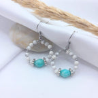 Paire de boucles d'oreilles créoles artisanales suspendues sur un tissu blanc. Elles sont composées de petites perles de howlite blanche marbrée de gris, avec une perle centrale facettée en amazonite bleu turquoise, encadrée par des intercalaires en argent.