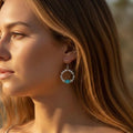 Gros plan de profil d'une femme portant une boucle d'oreille créole artisanale. Le bijou est composé d'un anneau de perles en howlite blanche et d'une perle centrale en amazonite turquoise. La scène est éclairée par une lumière naturelle chaude de fin de journée, mettant en valeur les reflets dorés de sa chevelure et les détails minéraux des pierres.