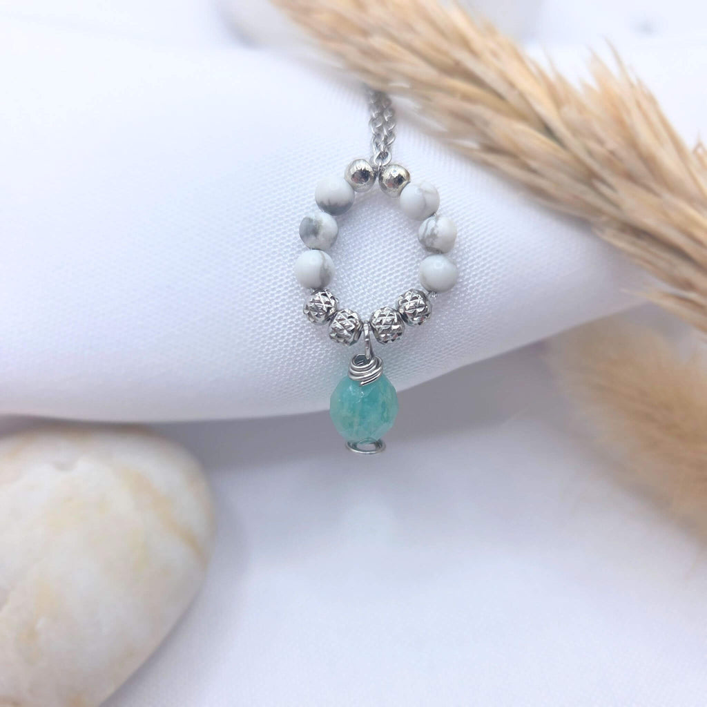 Pendentif artisanal composé d'un anneau de perles de Howlite blanche marbrée et de perles argentées, avec une goutte d'Amazonite facettée suspendue au centre, présenté sur un tissu blanc avec un épi de blé séché.
