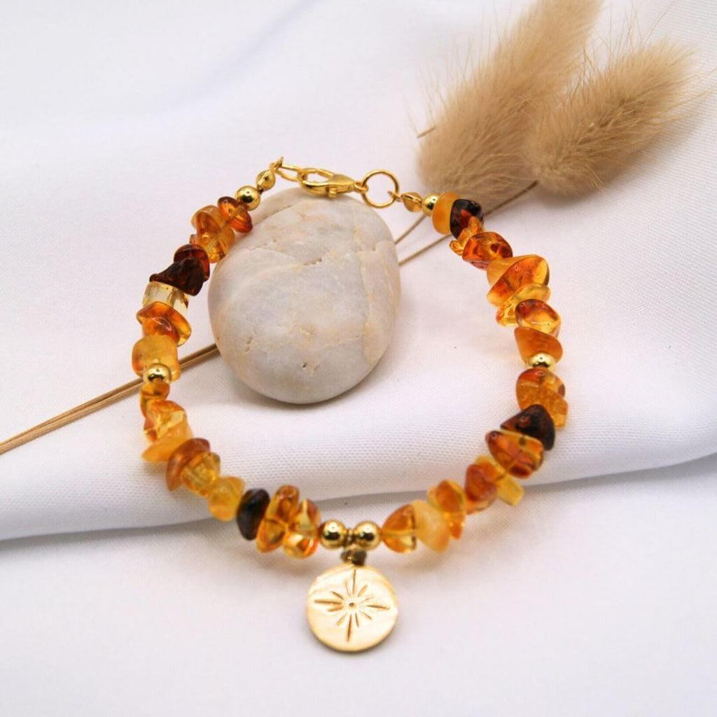 Un bracelet au style organique composé de perles d'ambre brut aux formes irrégulières et aux nuances allant du jaune miel au brun profond. Le bijou est agrémenté d'une petite médaille dorée circulaire gravée d'une étoile stylisée au centre. Présenté sur un tissu blanc drapé, il est accompagné d'un galet lisse et de fleurs séchées, créant une esthétique naturelle et solaire.