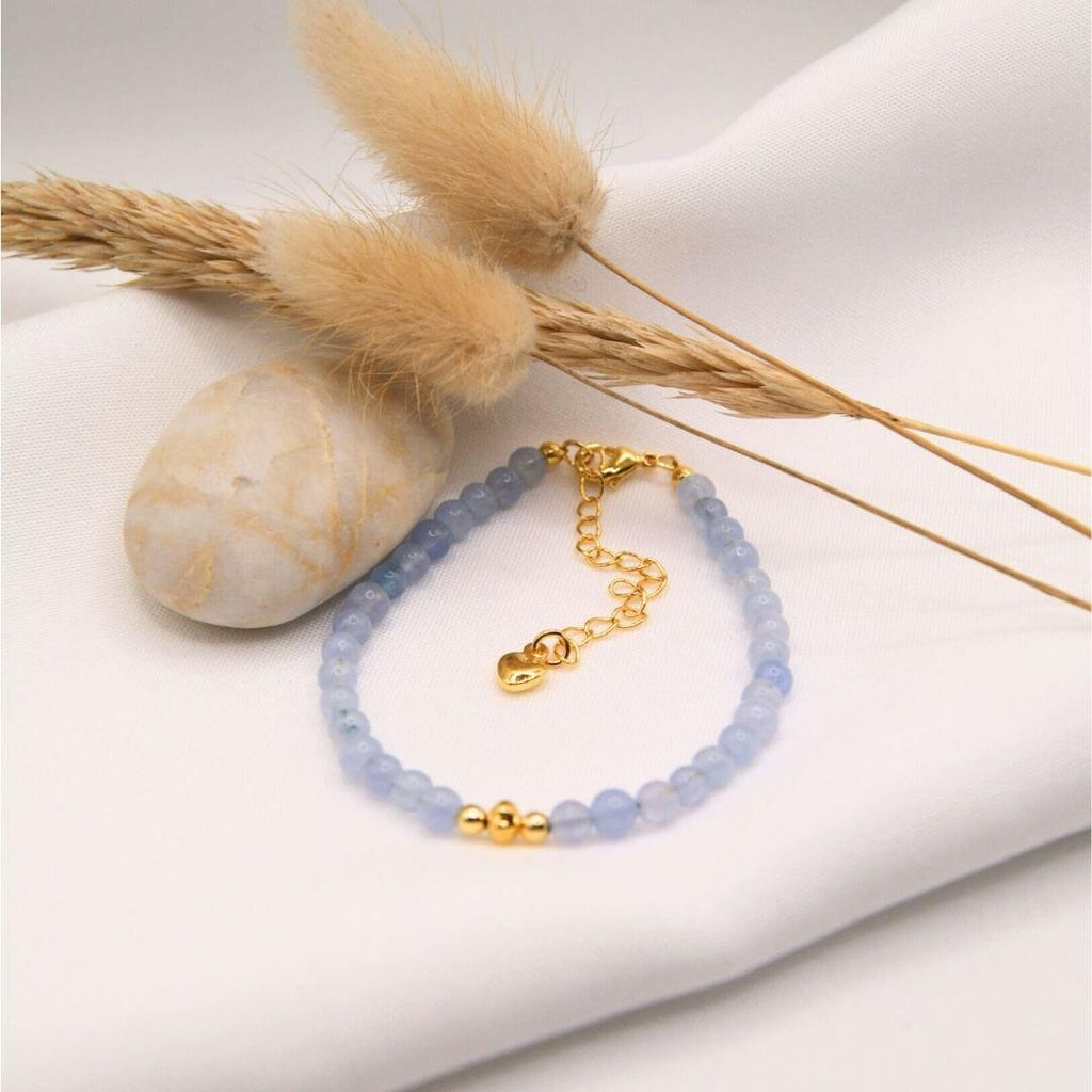 Un bracelet délicat composé de petites perles d'aigue marine bleue aux nuances douces et translucides. Le bijou est muni d'un fermoir mousqueton doré et d'une chaînette d'extension ornée d'un petit pendentif en forme de cœur. Trois perles dorées centrales apportent une touche de brillance au milieu des perles bleues. Le bracelet est mis en scène sur un tissu blanc avec un galet lisse et des éléments végétaux séchés, créant un univers apaisant et naturel.