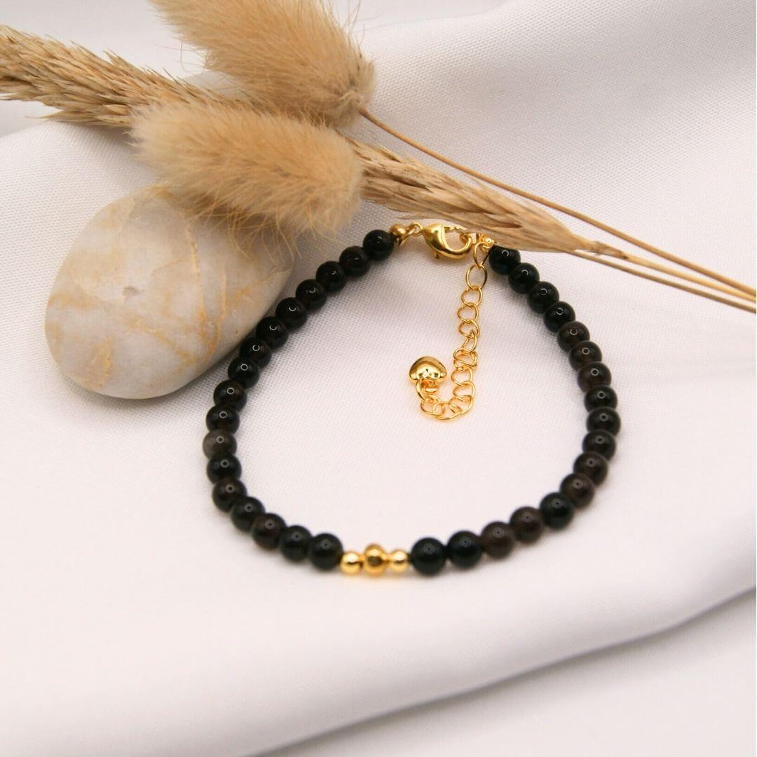 Un bracelet fin composé de petites perles d'onyx noires et brillantes. Le bijou dispose d'un fermoir mousqueton doré avec une chaînette d'extension terminée par un petit cœur. Au centre du bracelet, trois petites perles dorées sont regroupées pour créer un point focal. L'objet est mis en scène sur un tissu blanc avec un galet lisse, un épi de blé et des plumeaux séchés beiges.