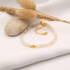 Un bracelet délicat composé de petites perles de pierre de lune aux reflets blancs laiteux et opalescents. Le bijou est doté d'un fermoir mousqueton doré et d'une chaînette d'extension agrémentée d'un petit pendentif en forme de cœur. Trois perles dorées lisses sont disposées au centre du bracelet. L'objet est présenté sur un tissu blanc avec un galet poli et des graminées séchées, évoquant une esthétique douce et pure.