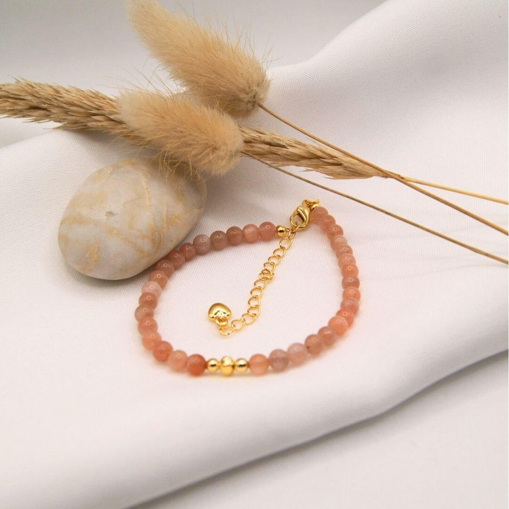 Un bracelet fin et élégant composé de petites perles de pierre de soleil aux nuances pêche et orangées. Le bijou dispose d'un fermoir mousqueton doré avec une chaînette d'extension terminée par un petit cœur. Au centre, trois perles dorées lisses créent un éclat chaleureux. L'ensemble est mis en scène sur un drapé de tissu blanc, accompagné d'un galet lisse et de graminées séchées.