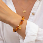 Gros plan sur le poignet d'une personne portant un bracelet en éclats d'ambre aux nuances chaudes de miel et de cognac. Le bijou est orné d'une petite médaille ronde dorée gravée d'une étoile rayonnante.